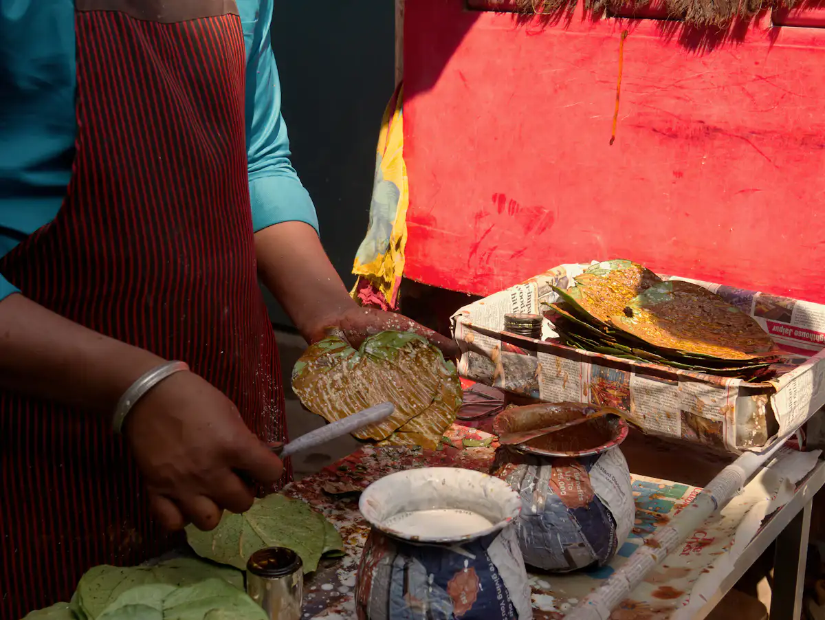 A person in a striped apron spreading paste onto betel leaves at a street stall — The paan and digestive cart