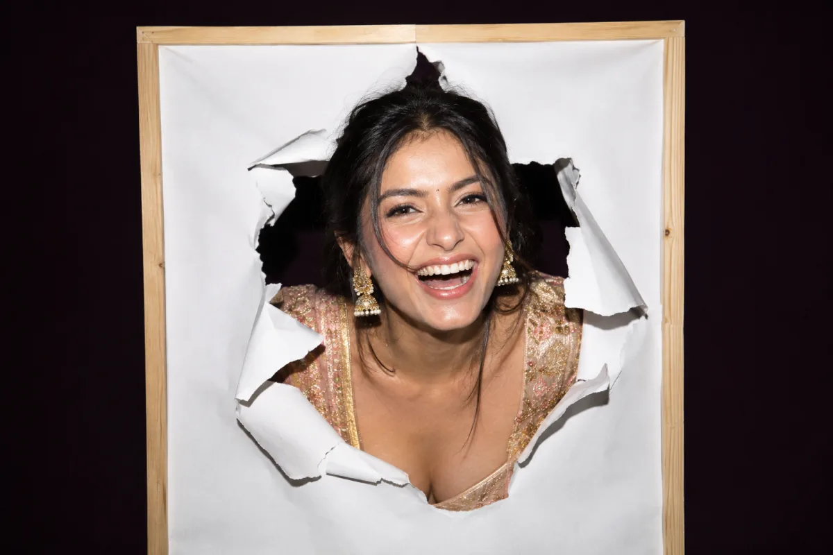 A laughing woman in a gold and rose lehenga bursting head-first through a torn sheet of white paper in a wooden frame, lit by hard flash — The paper wall guests punch their face through